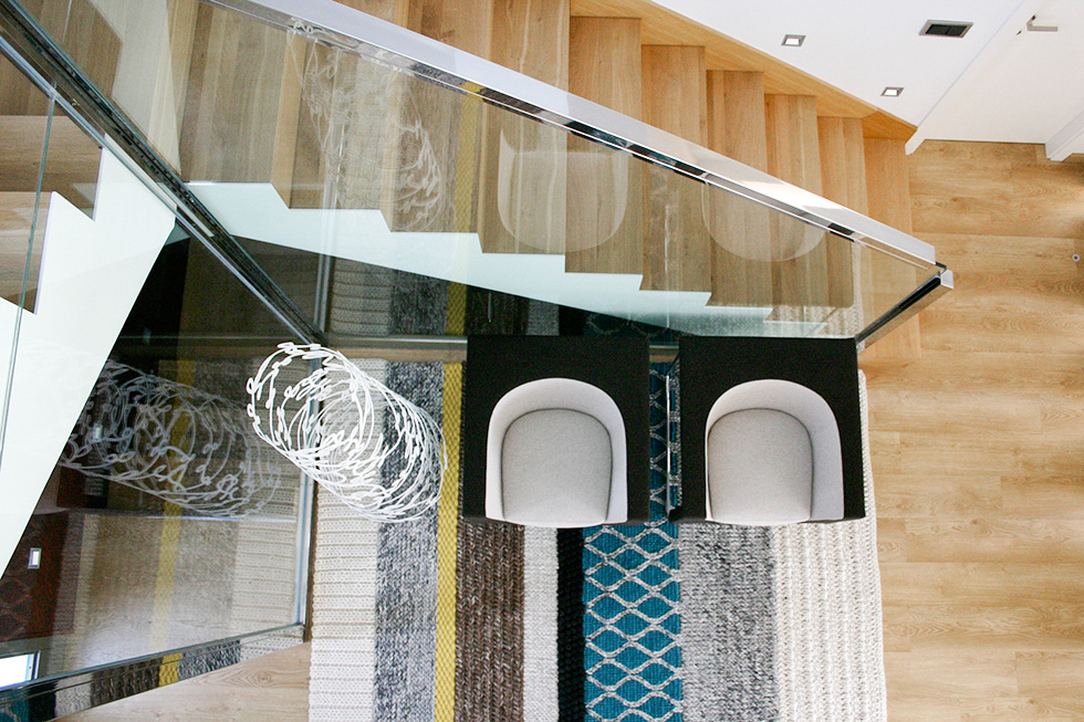 Recibidor moderno con escalera de madera y barandilla de acero y cristal, con sillones Stua y alfombra Gan.